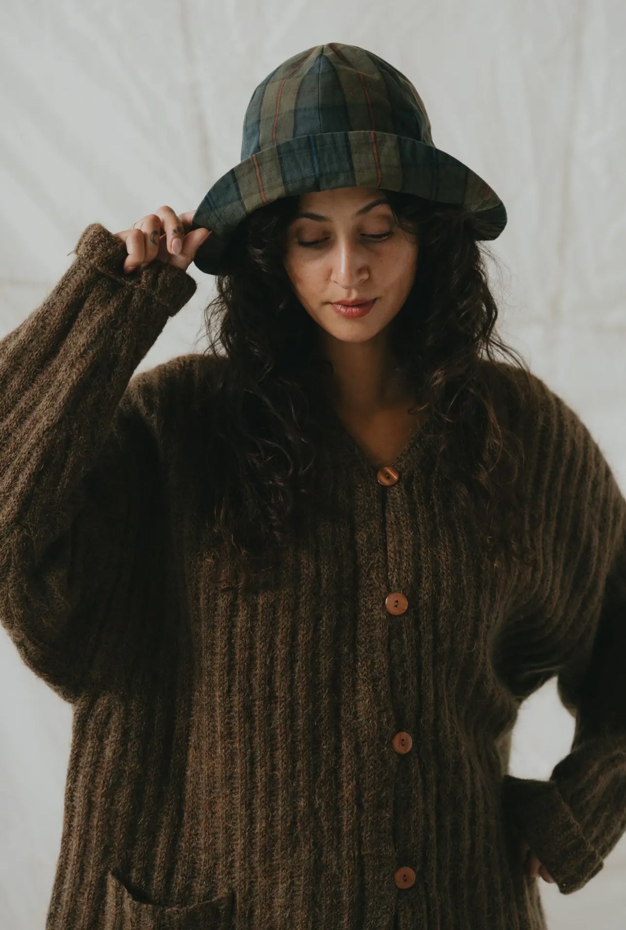 Woman wearing a brown knitted cardigan and plaid hat against a white background
